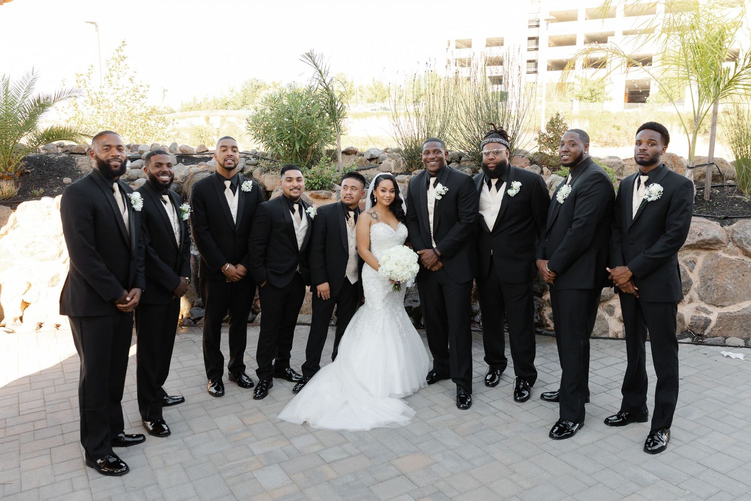 Wedding party in formal attire poses outdoors for group photos at a scenic venue.