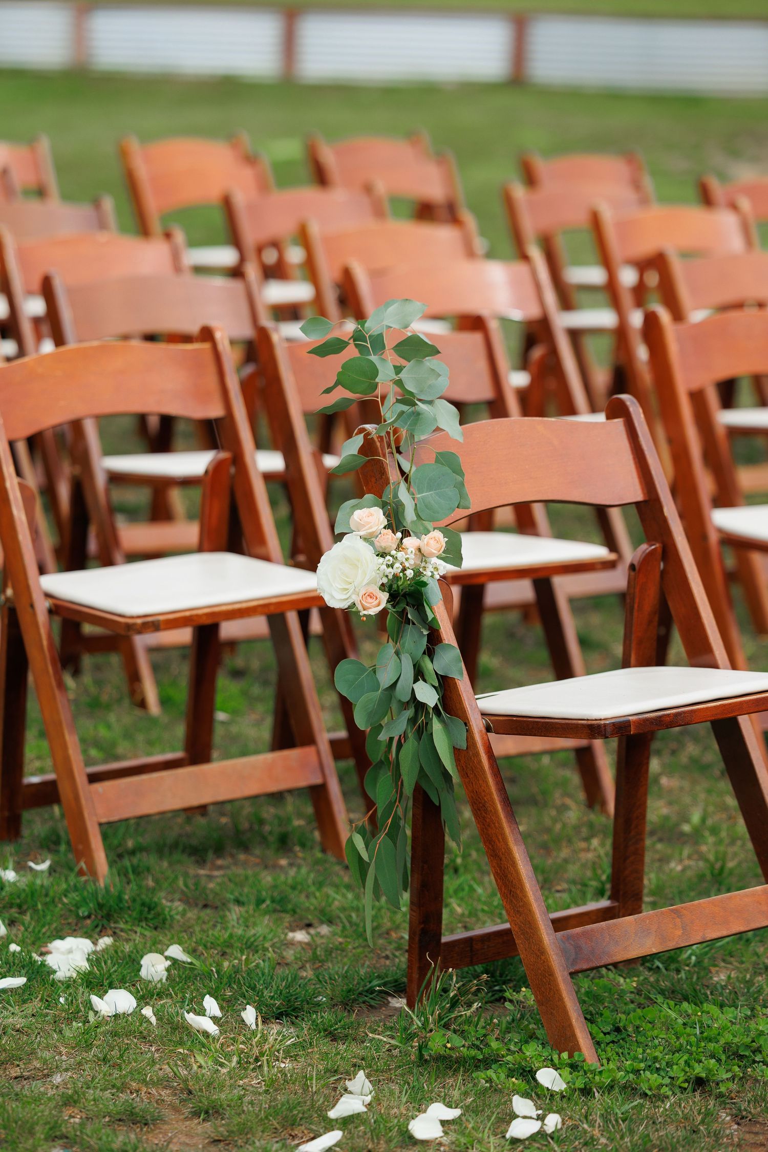 Wooden folding chairs decorated with white flowers and eucalyptus leaves arranged on grass for an outdoor wedding ceremony.