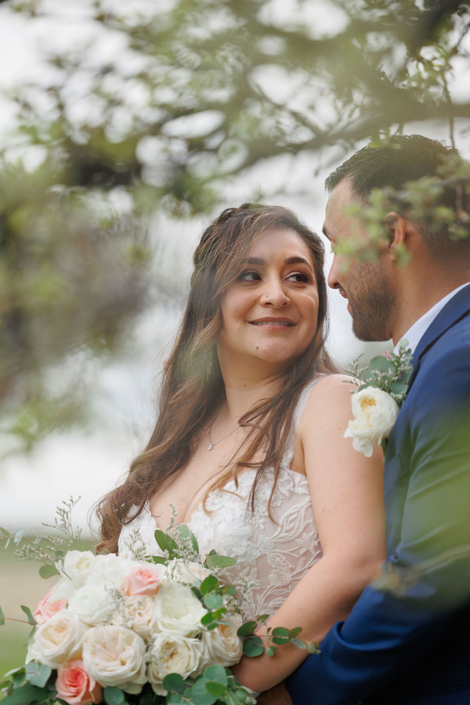 A romantic wedding couple embraces outdoors with soft pink roses and greenery in a dreamy garden setting.