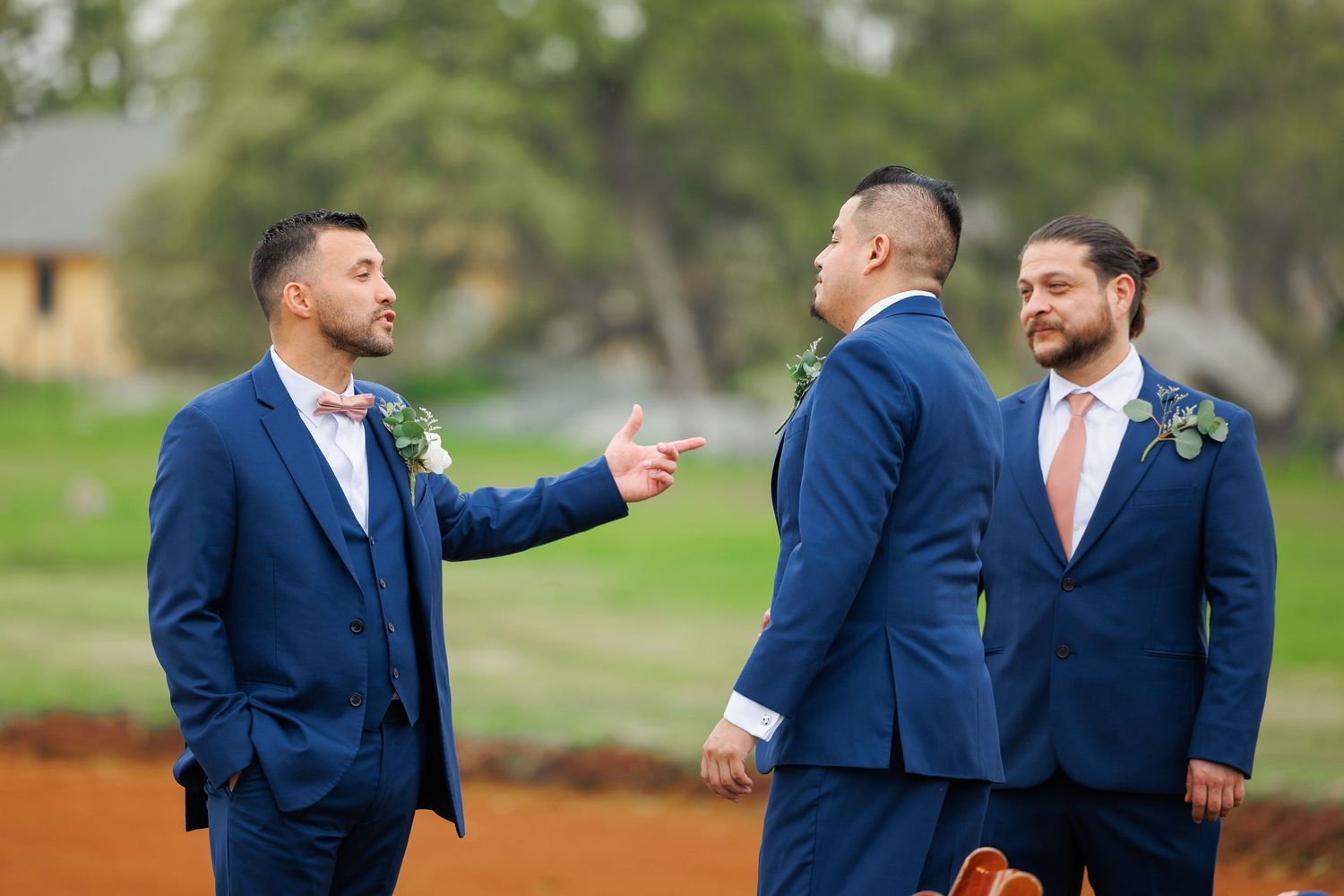 Groomsmen in navy blue suits share a candid moment of laughter and conversation during an outdoor wedding.