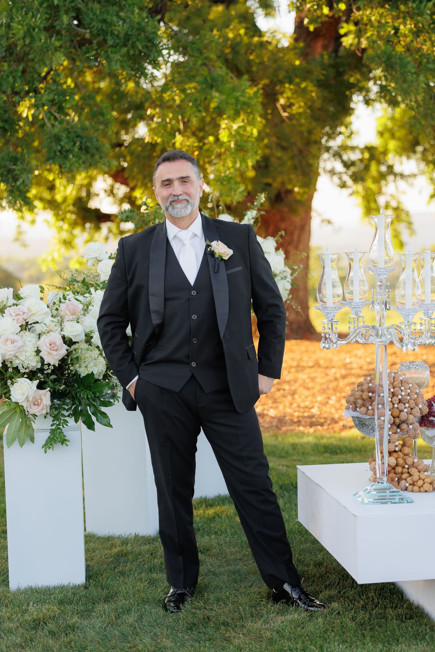 Wedding reception setup outdoors with white benches and floral arrangements at sunset in a garden setting.