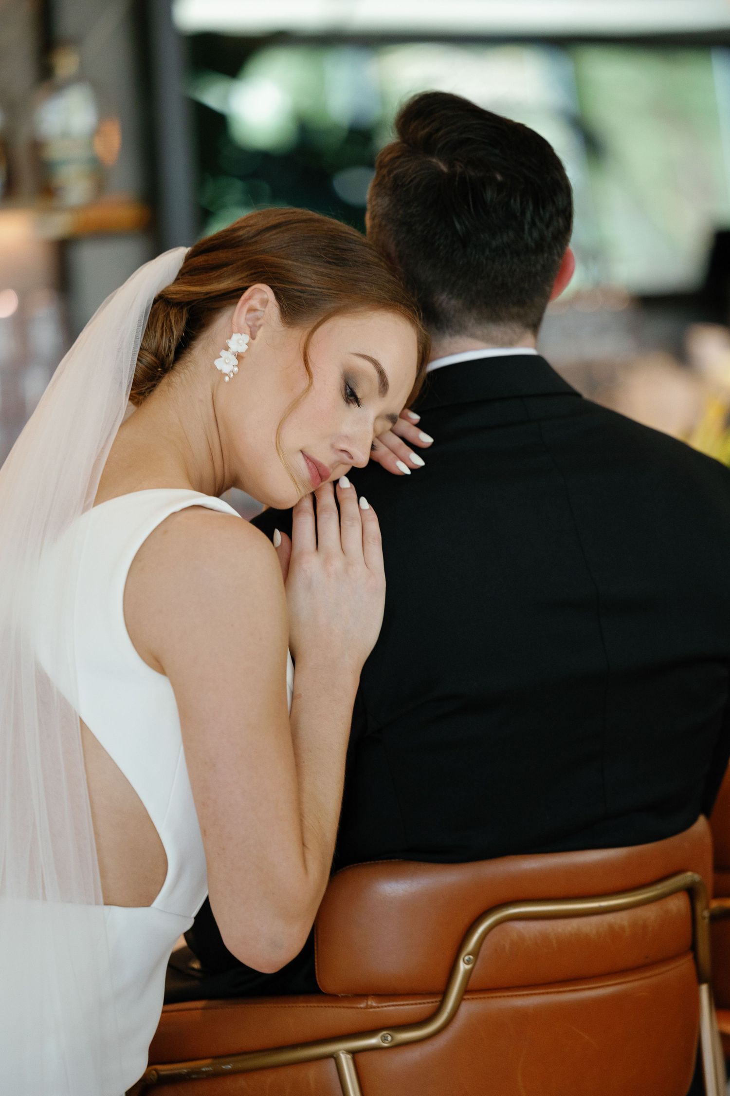 Bride in white dress rests her head on groom's shoulder while seated in brown leather chairs at wedding reception.