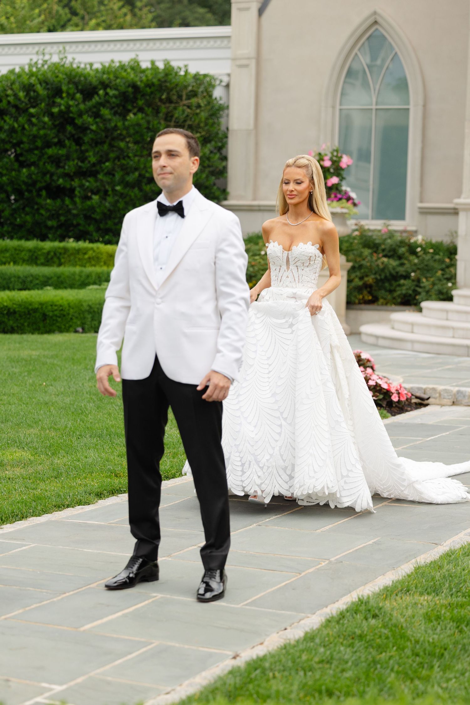 A bride and groom walk together on a stone path outside a white church with Gothic windows and lush greenery.