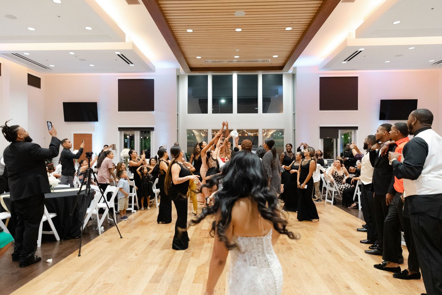 Wedding reception guests celebrate on dance floor as newlywed couple dances in modern venue with wood floors and white decor.