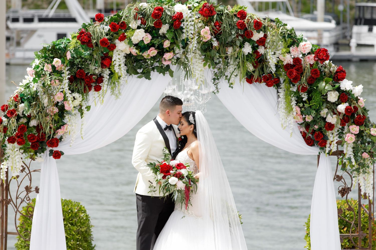 A wedding ceremony setup with a floral arch decorated in red and white roses overlooking a waterfront.