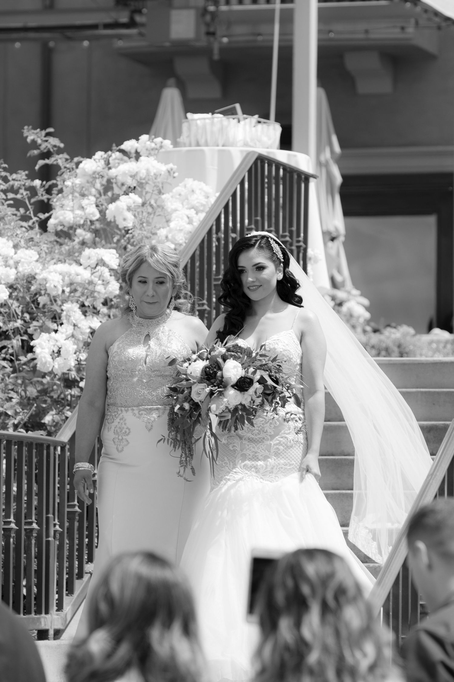 Black and white wedding photo showing two people in formal attire standing together outdoors with flowers and railings.