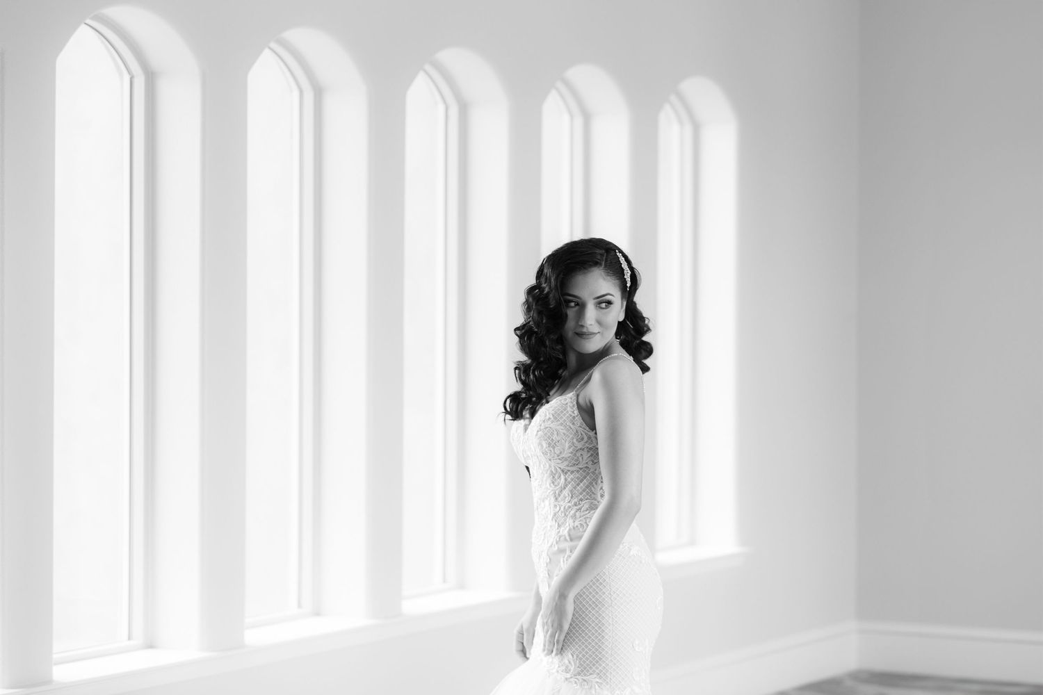 A bride in a white wedding dress stands in a bright sunlit room with tall windows creating an ethereal atmosphere.