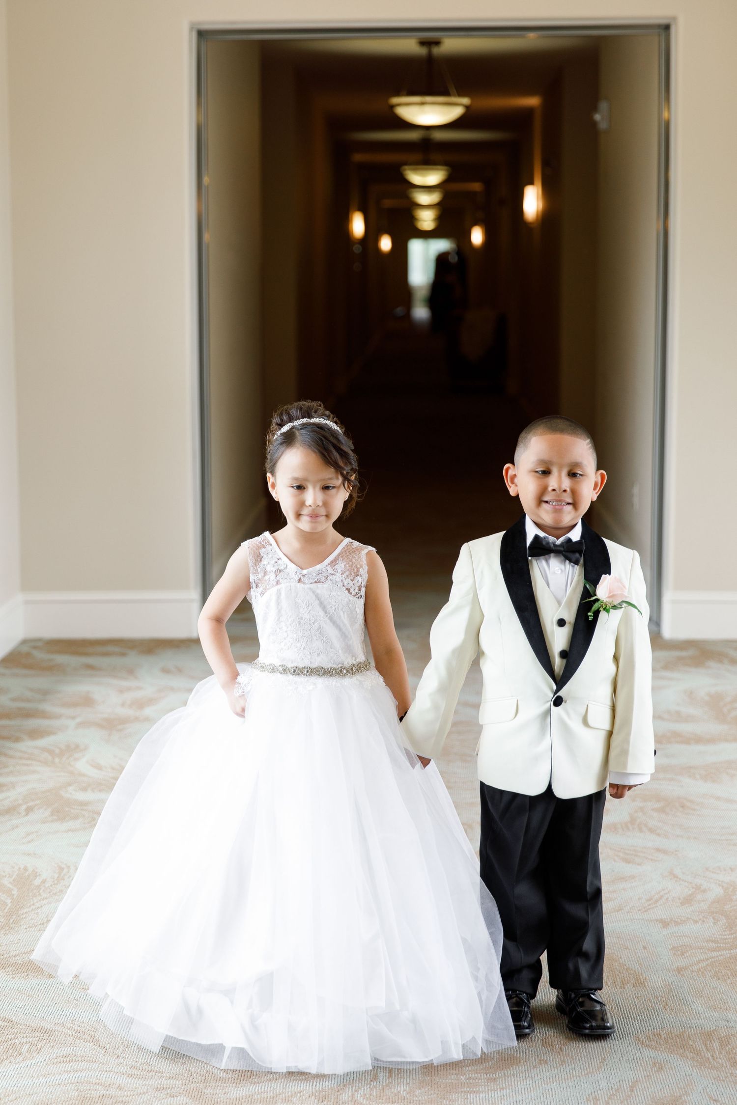 Two young children dressed in formal white wedding attire stand together in an elegant hallway with pendant lighting.