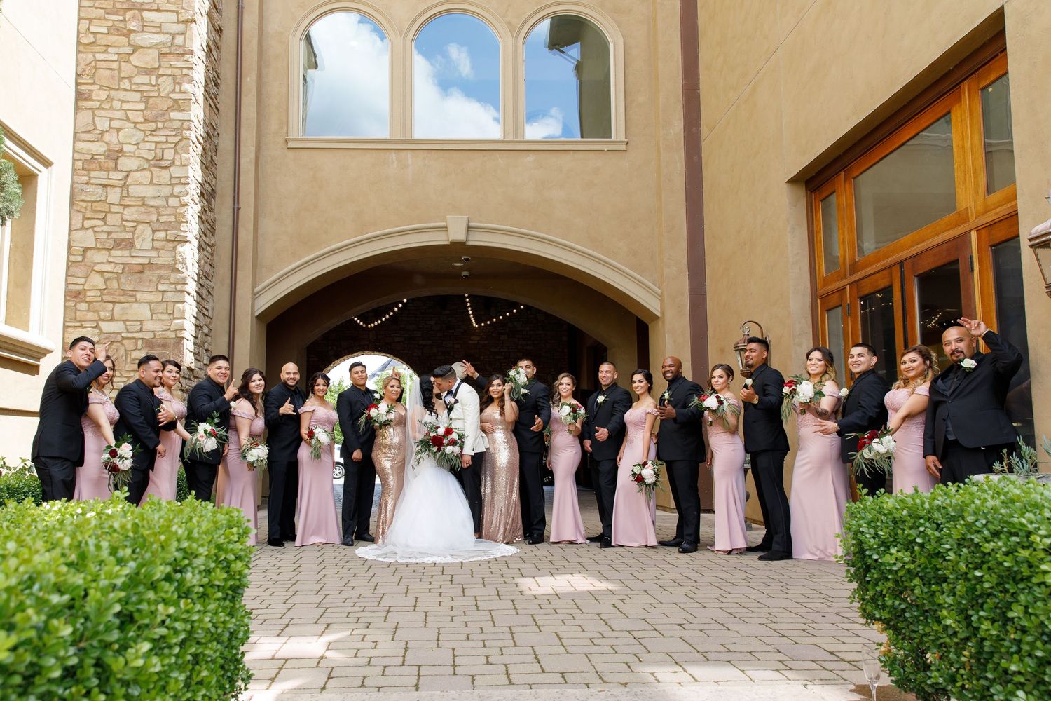 Wedding party in pink dresses poses for a panoramic photo outside a stone church entrance.