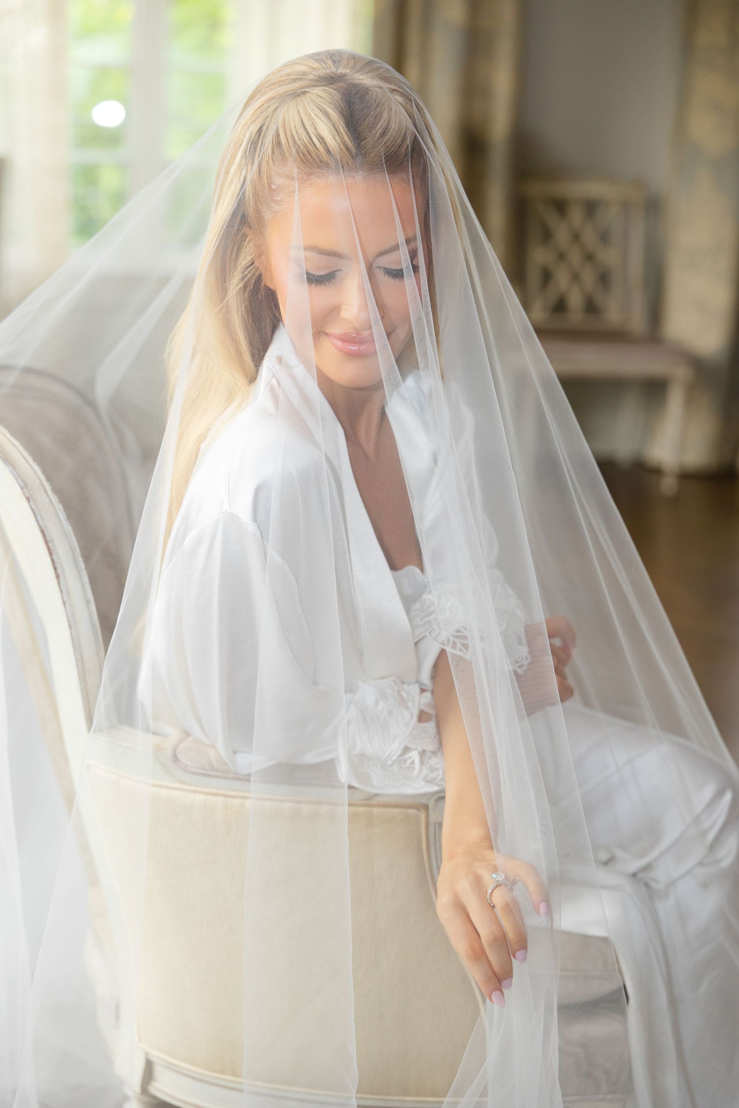 Bride in white robe and veil sits gracefully on elegant chair, smiling softly in bright bridal suite.