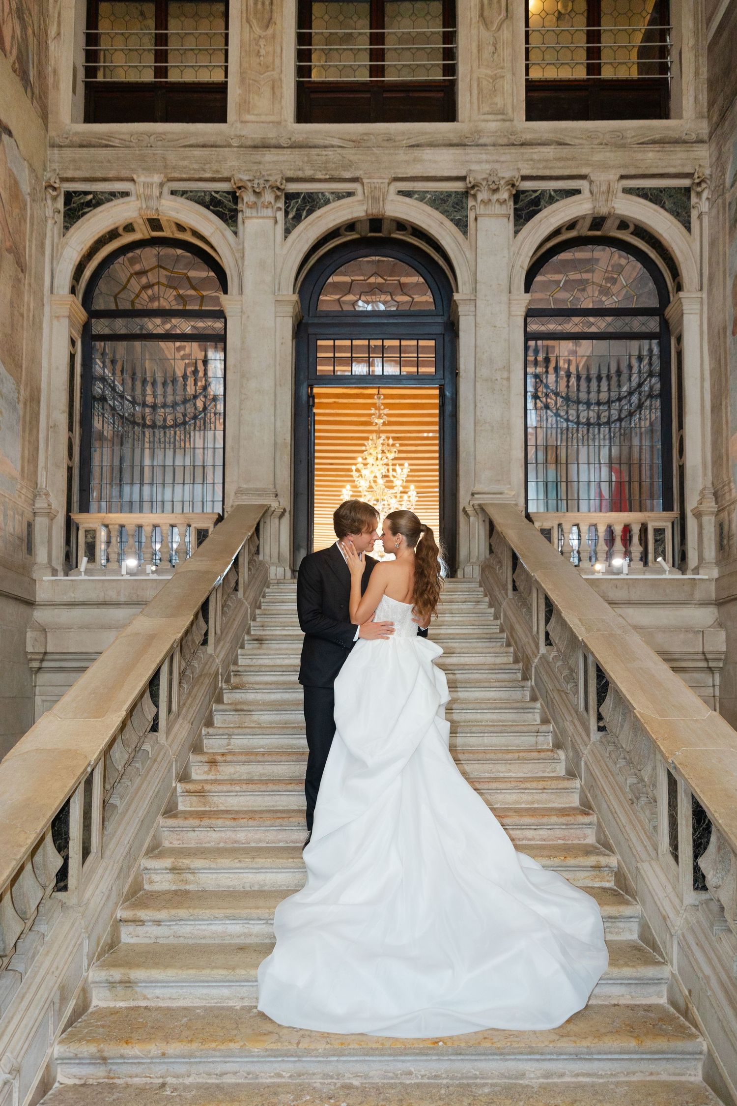 A couple embraces on an elegant marble staircase with ornate arched windows and architectural details in a grand building.