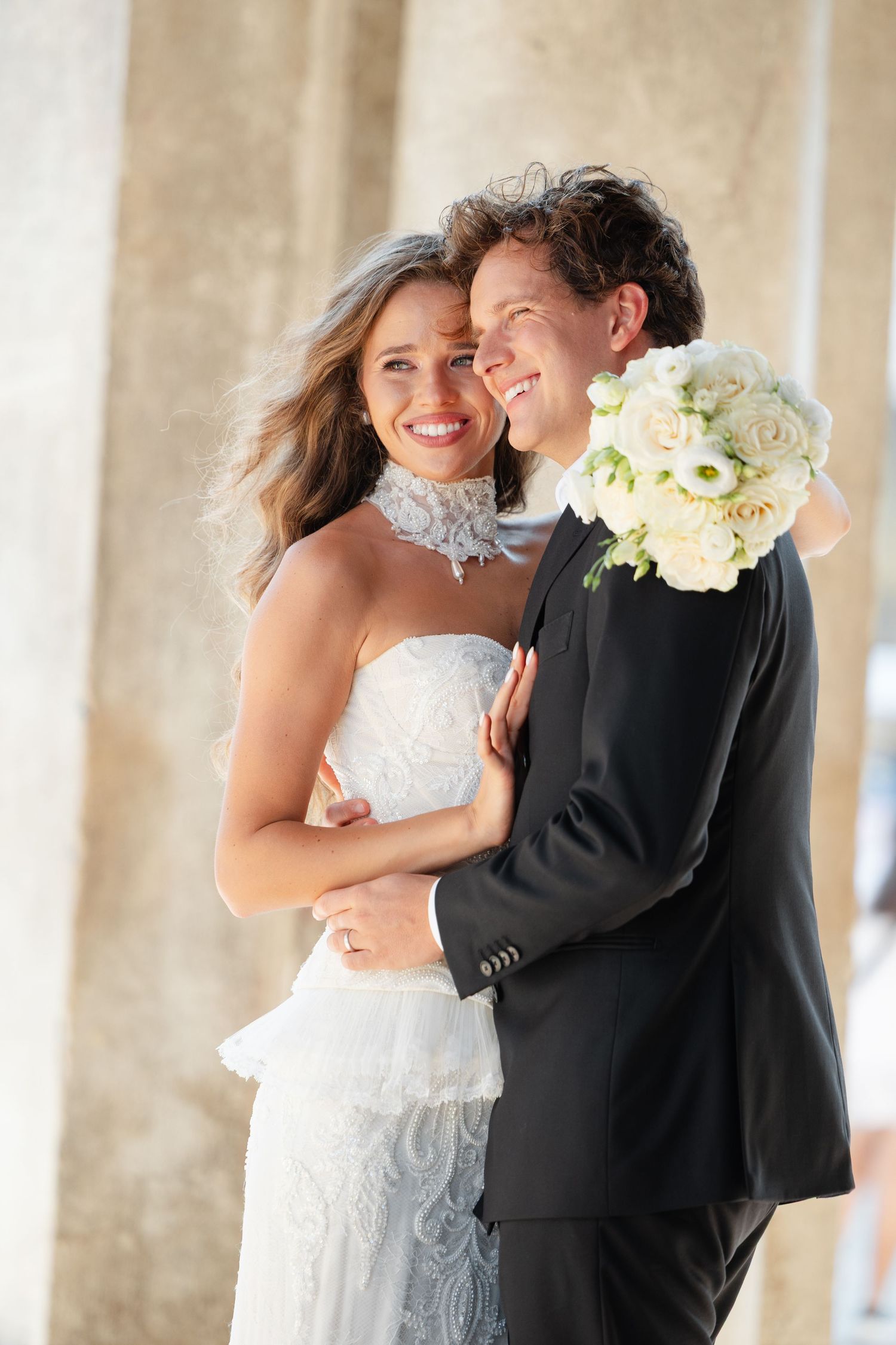 A happy couple embraces on their wedding day, with the bride holding a white rose bouquet and wearing a lace gown.