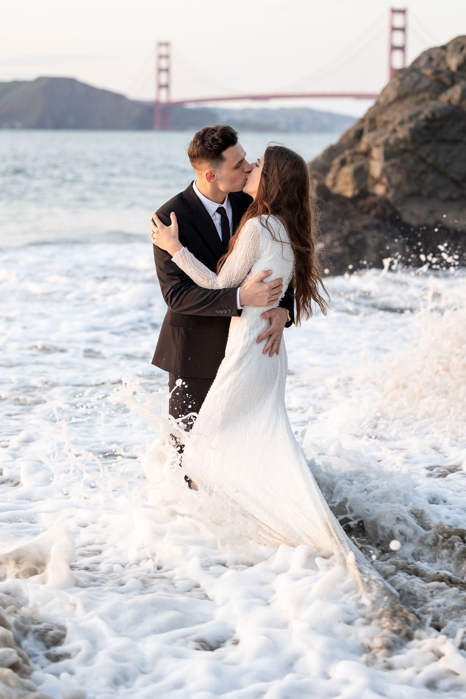 Romantic couple embracing on rocky shore with Golden Gate Bridge and crashing waves in background.