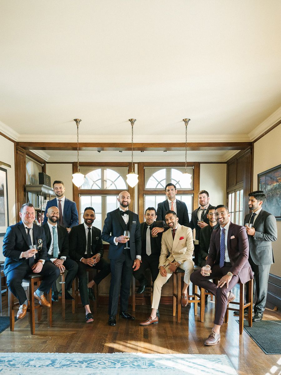 Groomsmen in formal suits pose together on wooden chairs in an elegant indoor setting.