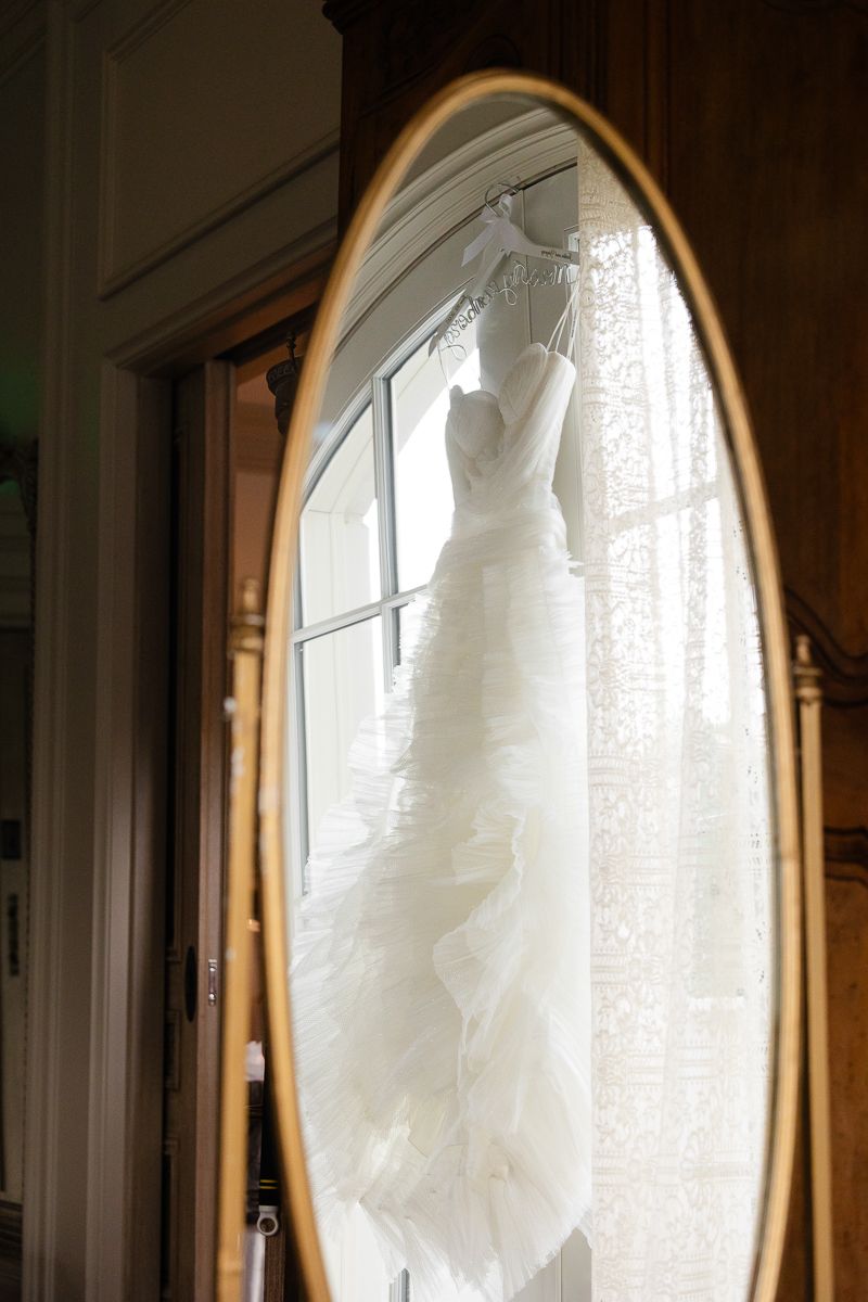 A white wedding dress hangs in an arched doorway, backlit by natural light creating a romantic silhouette.