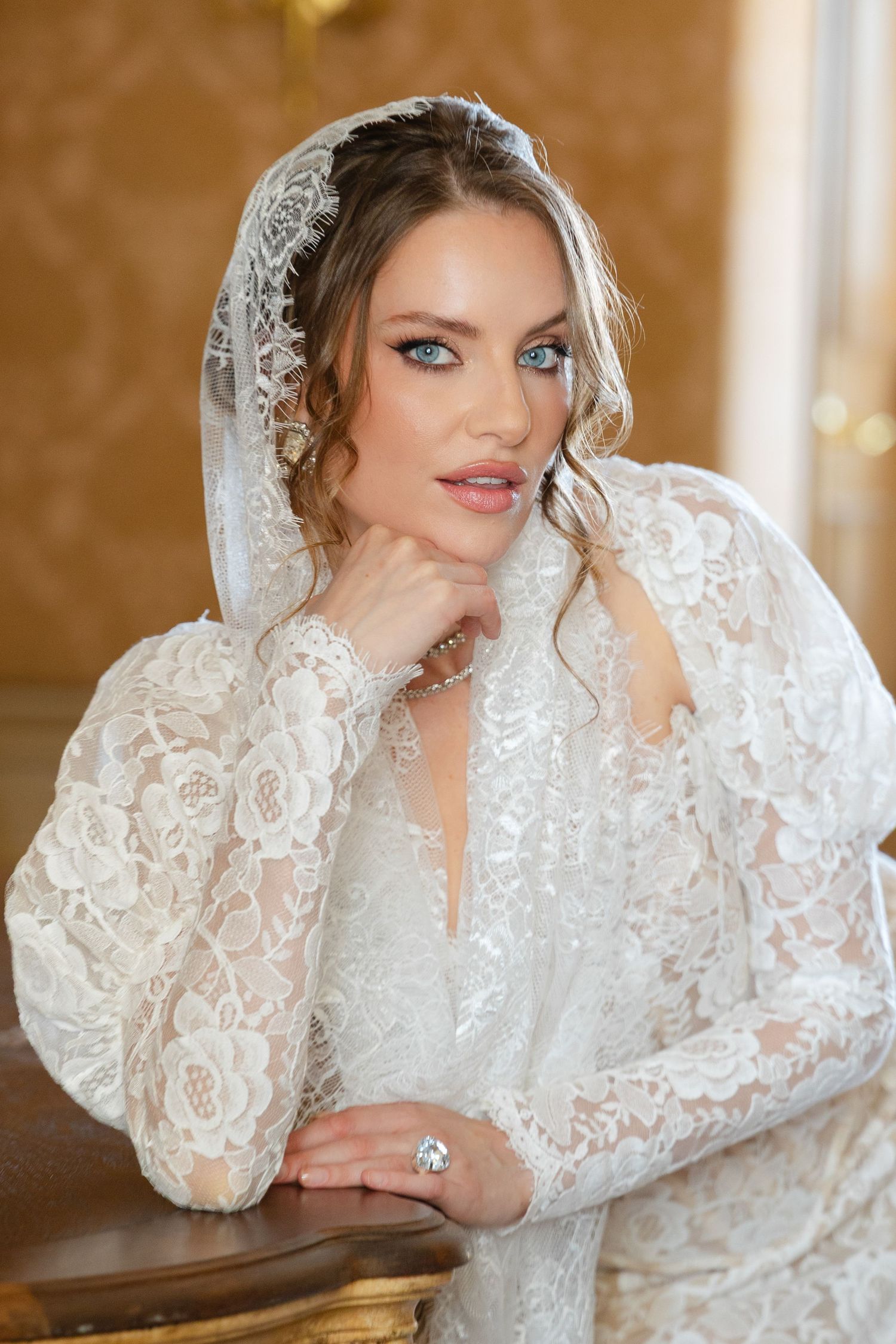 A bride in an elegant white lace wedding dress with long sleeves and veil poses against a golden-toned background.