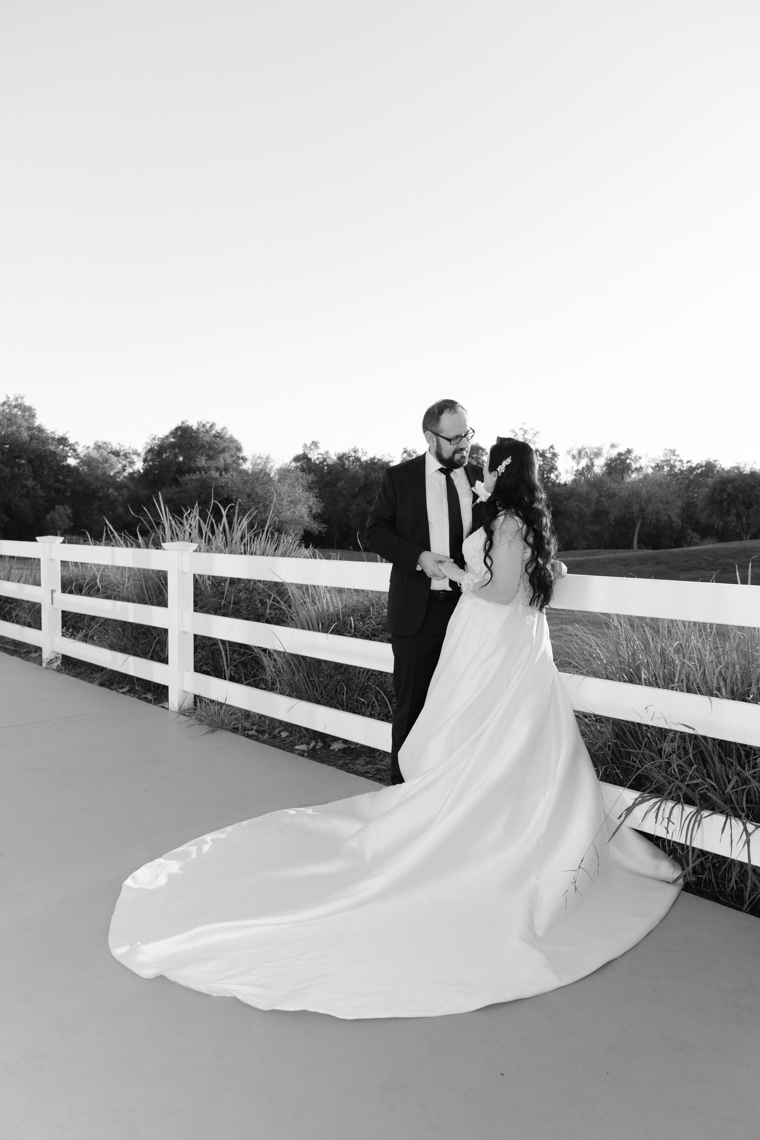 Black and white wedding photos by a white fence showing flowing bridal gown.