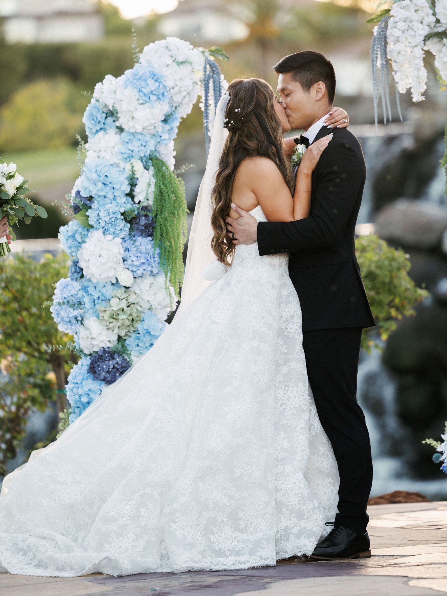 Newlyweds share a romantic kiss under an arch decorated with blue hydrangeas and white flowers.