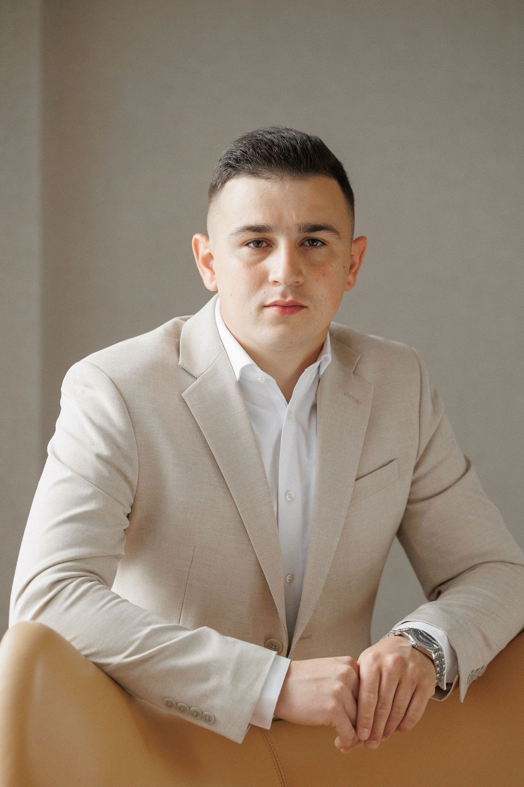 Professional portrait of someone wearing a beige suit jacket and white shirt in a minimalist setting.