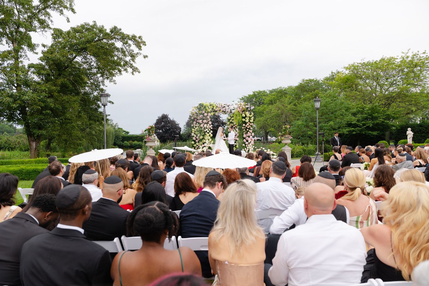 Wedding guests seated outdoors watching ceremony at a garden venue with trees in the background.
