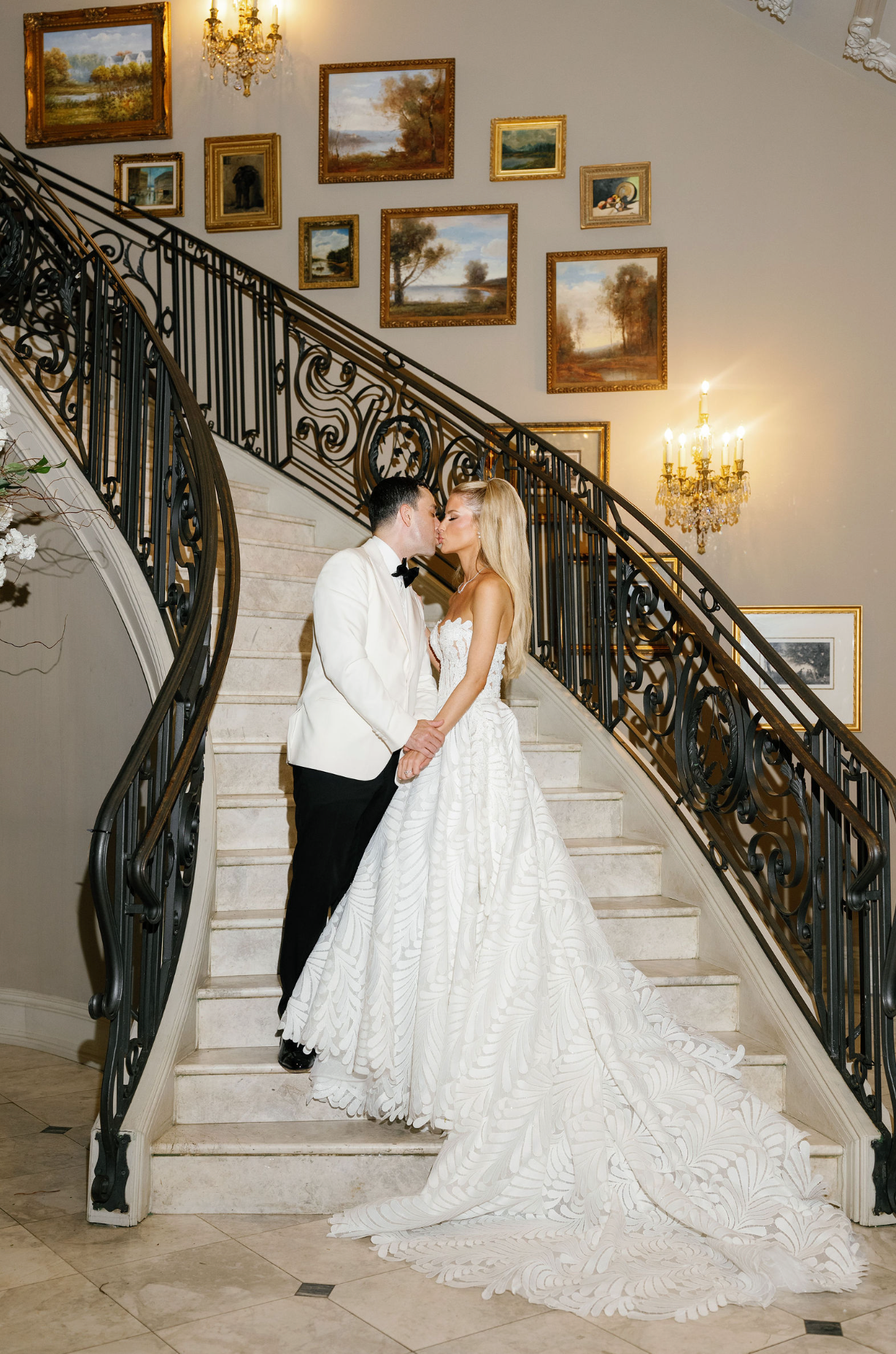 Romantic wedding couple shares intimate kisses on an elegant staircase with ornate railings and framed artwork on walls.
