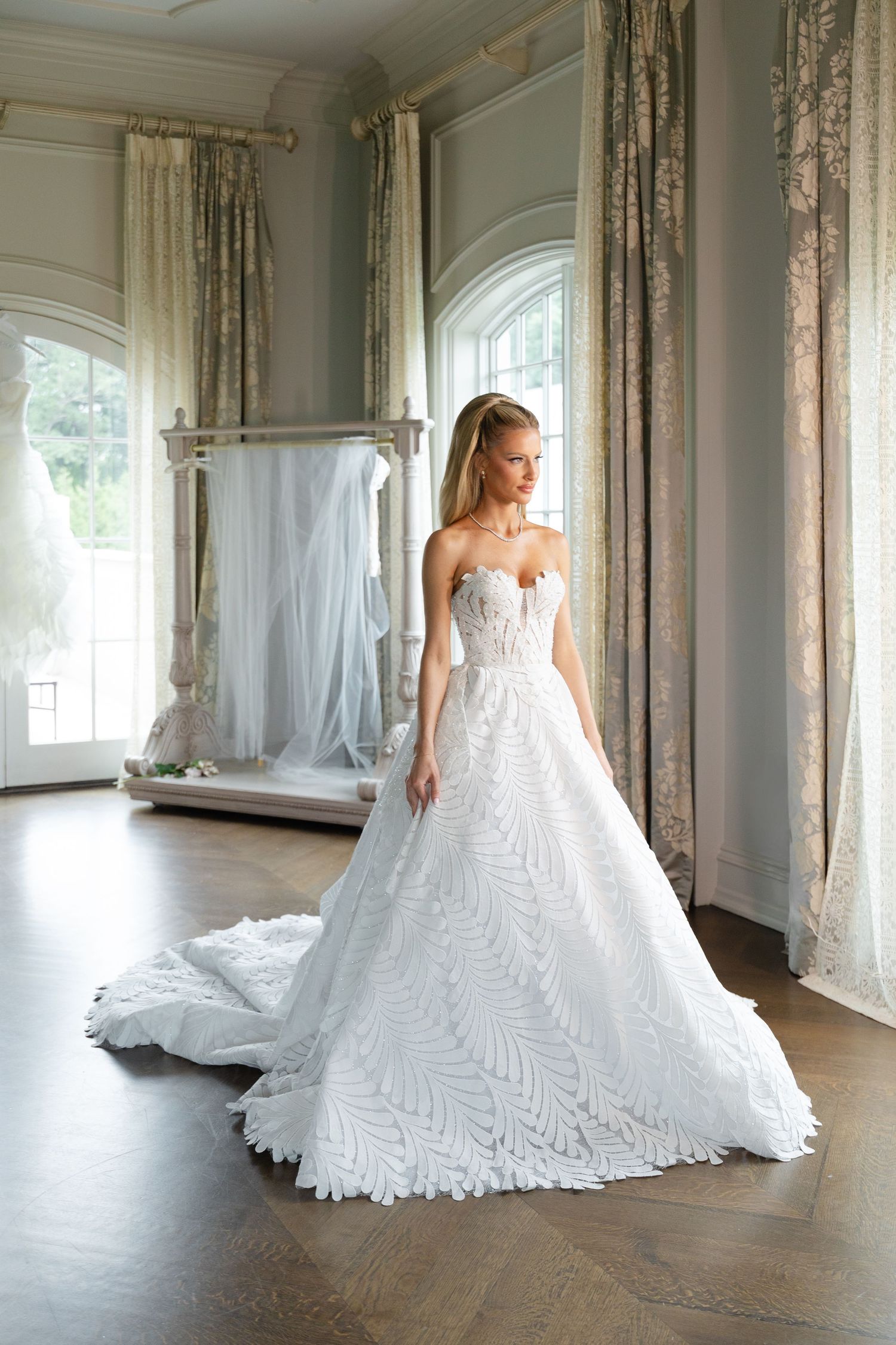 Elegant white wedding gown with intricate lace patterns and flowing train displayed in a bright, classic interior room.