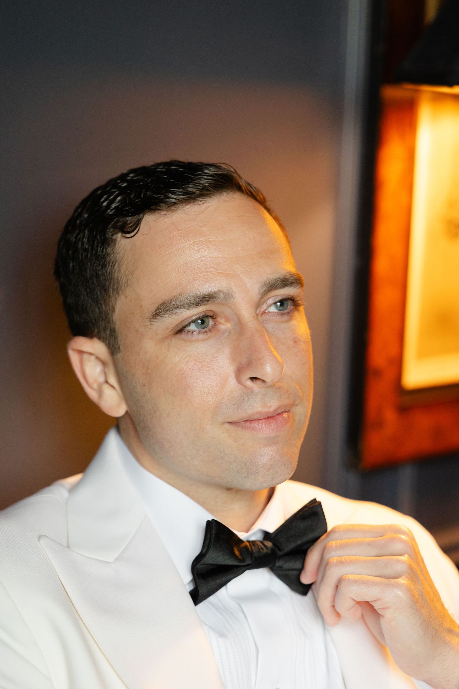 A person in a white dress shirt and black bowtie adjusts their collar in a dimly lit room.