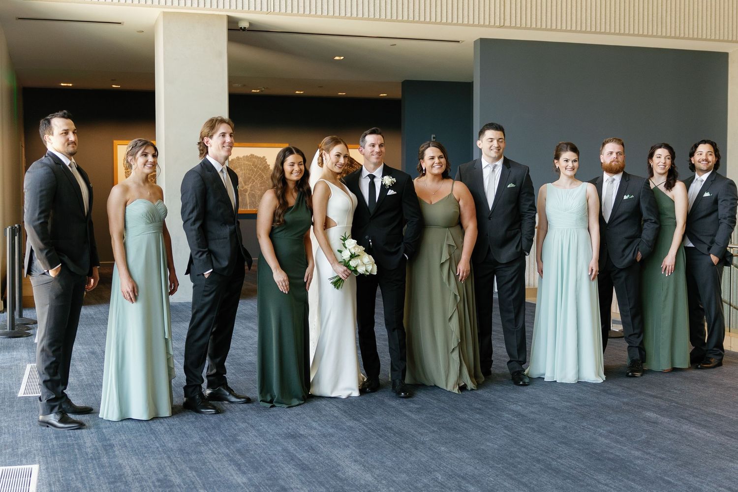 Wedding party members in formal attire line up against a beige wall in alternating light blue dresses and dark suits.