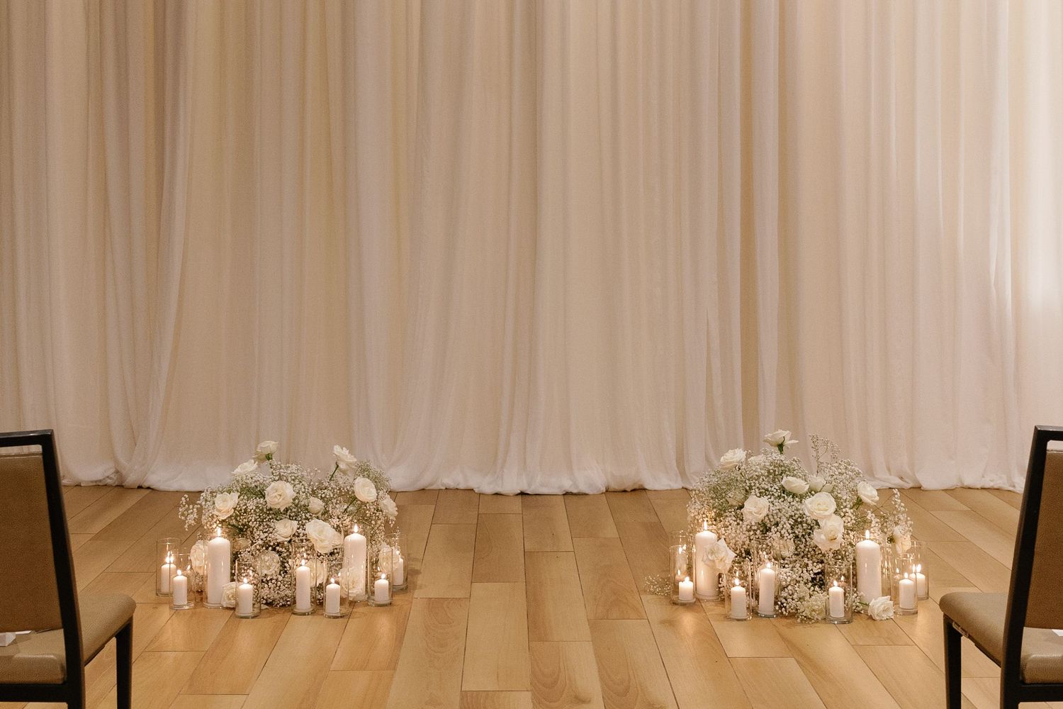 White floral centerpieces with candles line a long dining table with wooden chairs in an elegant reception space.