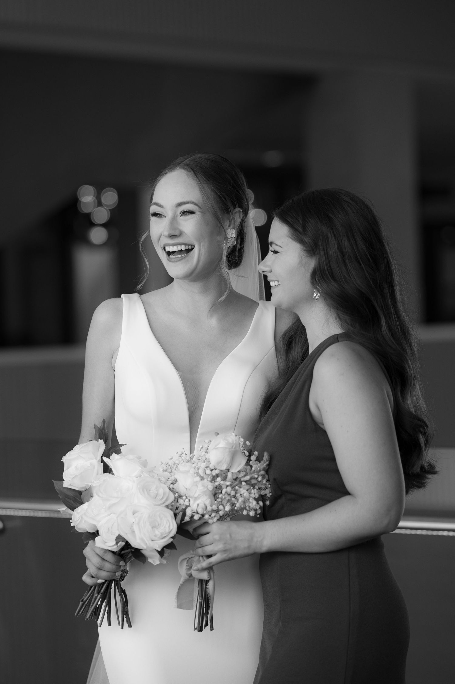 Black and white candid photo showing bridesmaids in formal dresses sharing joyful laughter while holding bouquets.