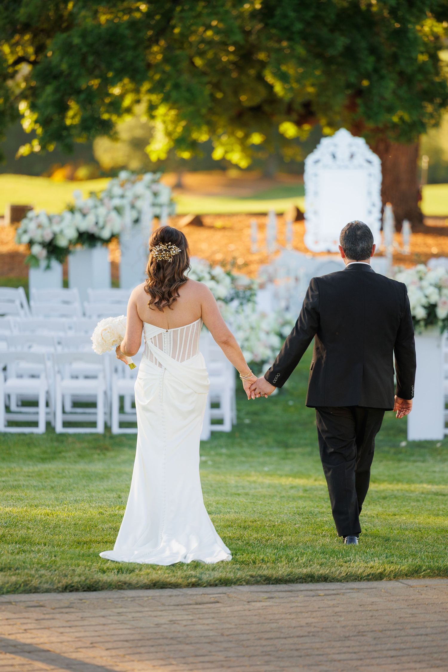A couple in wedding attire walks away from white chairs set up for an outdoor garden ceremony at sunset.