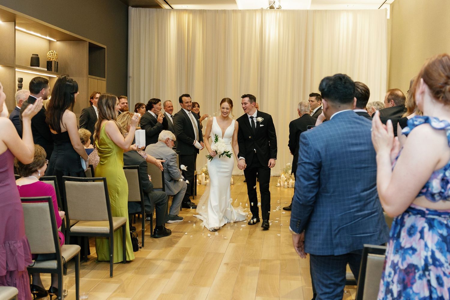 Wedding ceremony taking place in a modern venue with wooden floors and guests seated on both sides of the aisle.