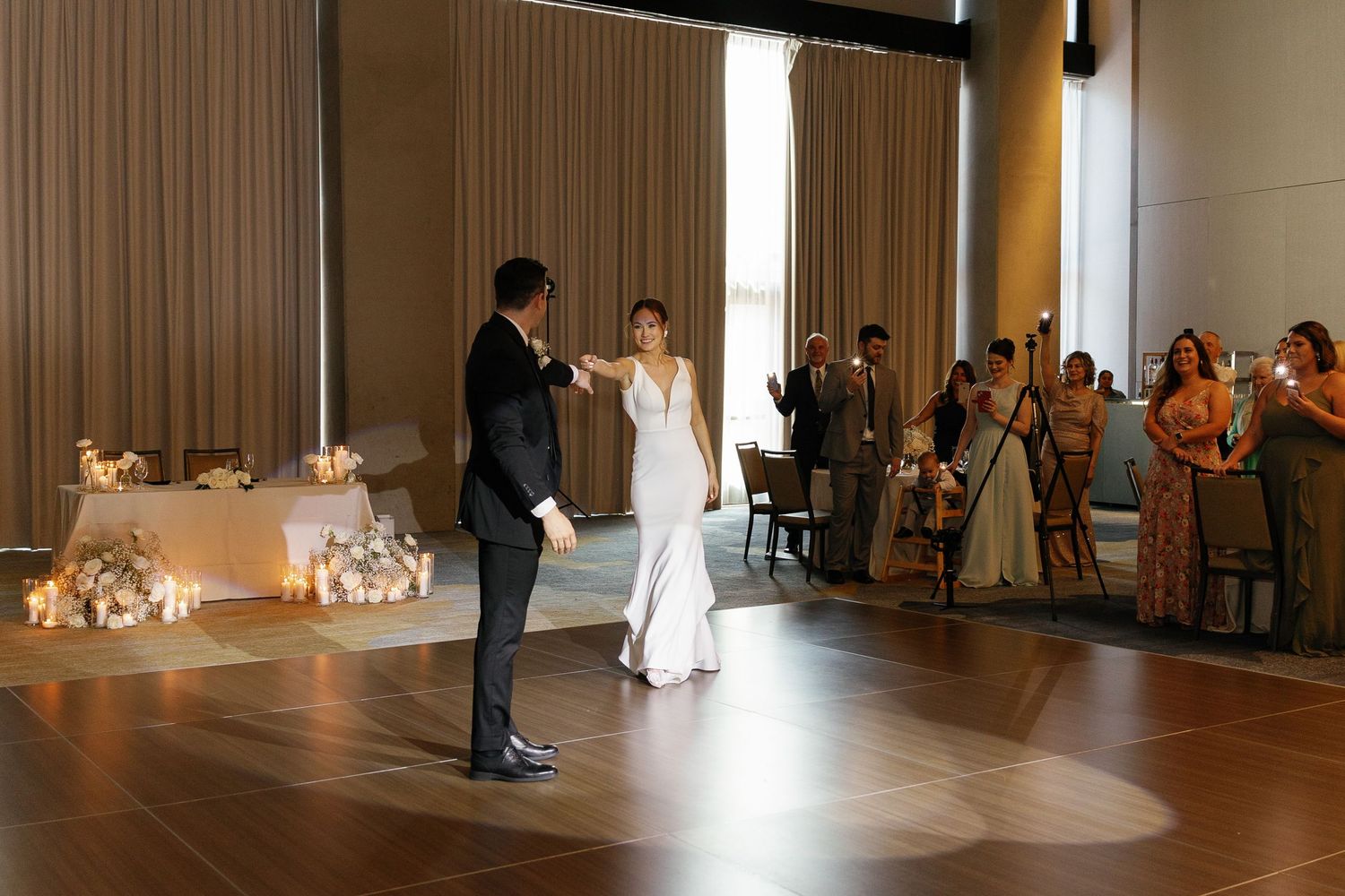 A couple shares their first dance at a modern reception venue with guests watching from the side and floral arrangements nearby.