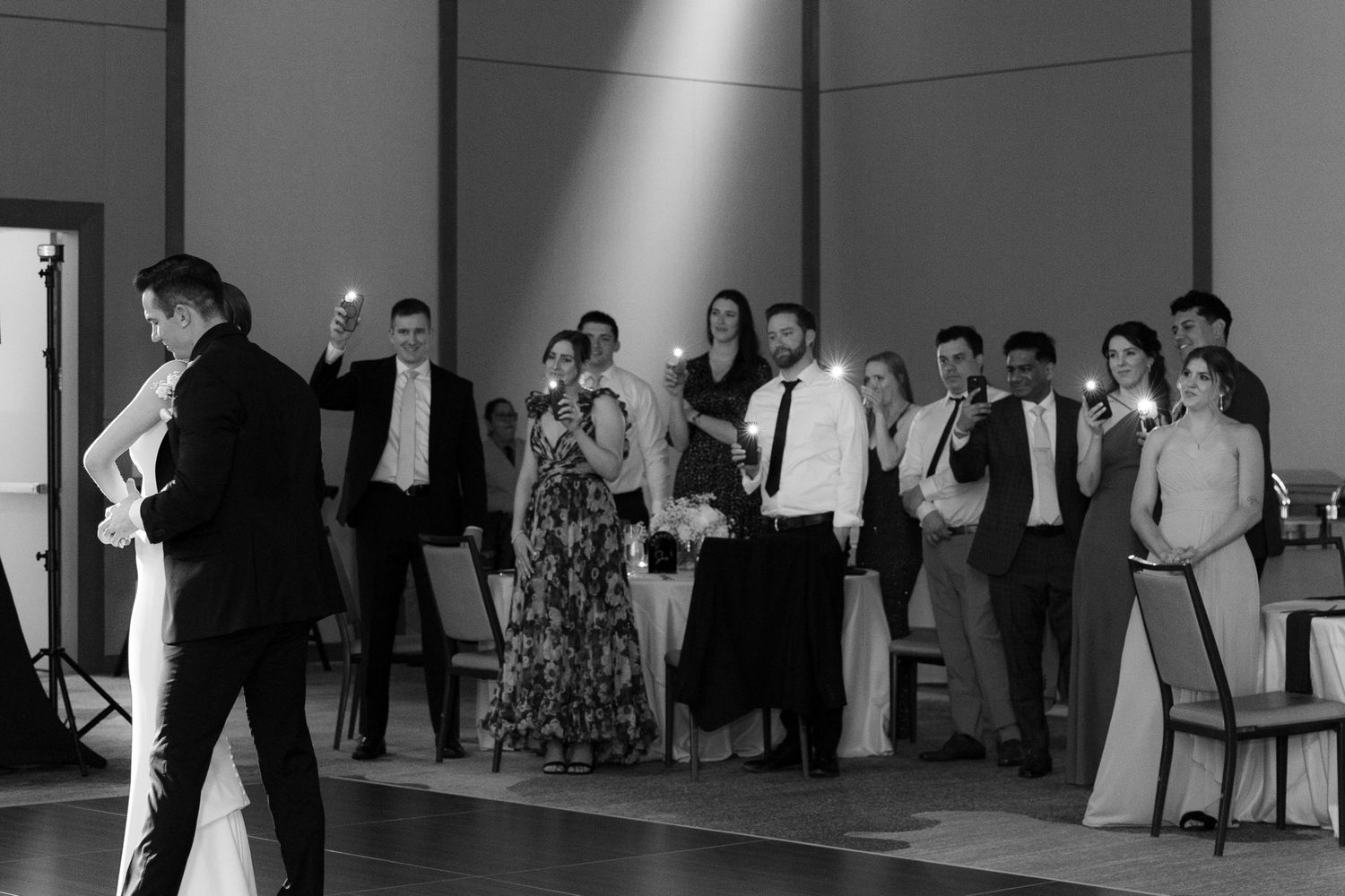 Wedding guests watch a couple dance during their first dance at a modern venue with white walls and dark floors.
