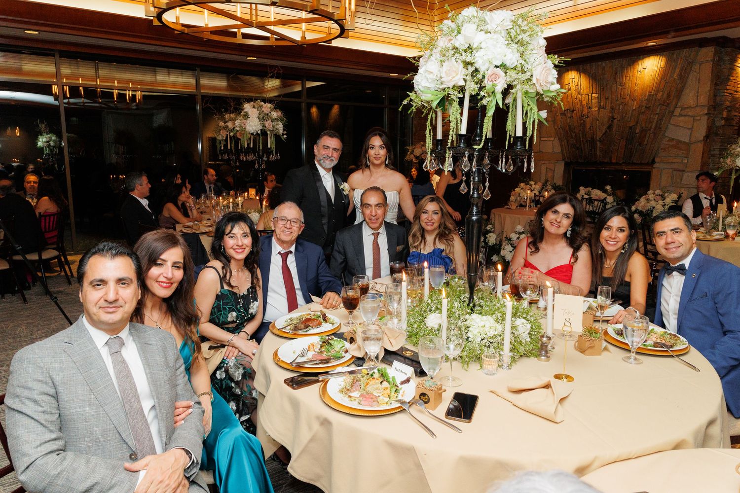 Wedding reception guests seated at elegant tables with floral centerpieces in a warmly lit banquet hall.