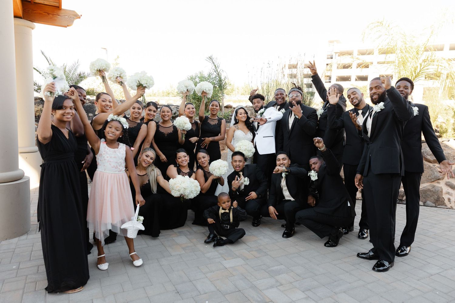 A large wedding party dressed in formal black and blush pink attire poses together outdoors at a venue.