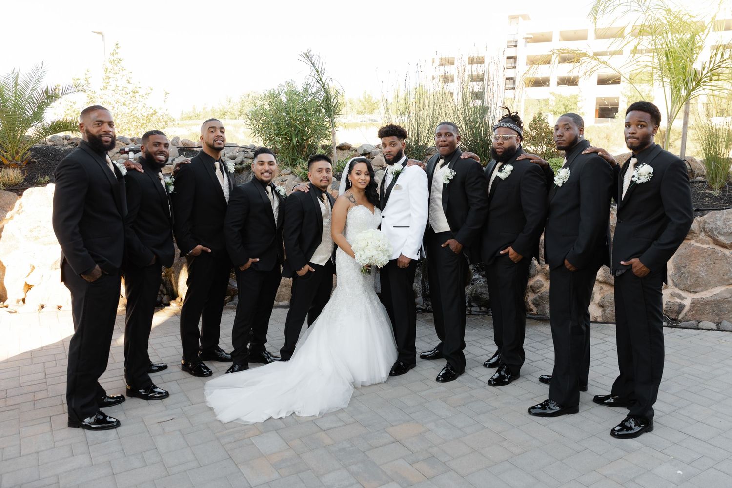 Wedding party poses outdoors in formal black and white attire on a sunny day at an elegant venue.