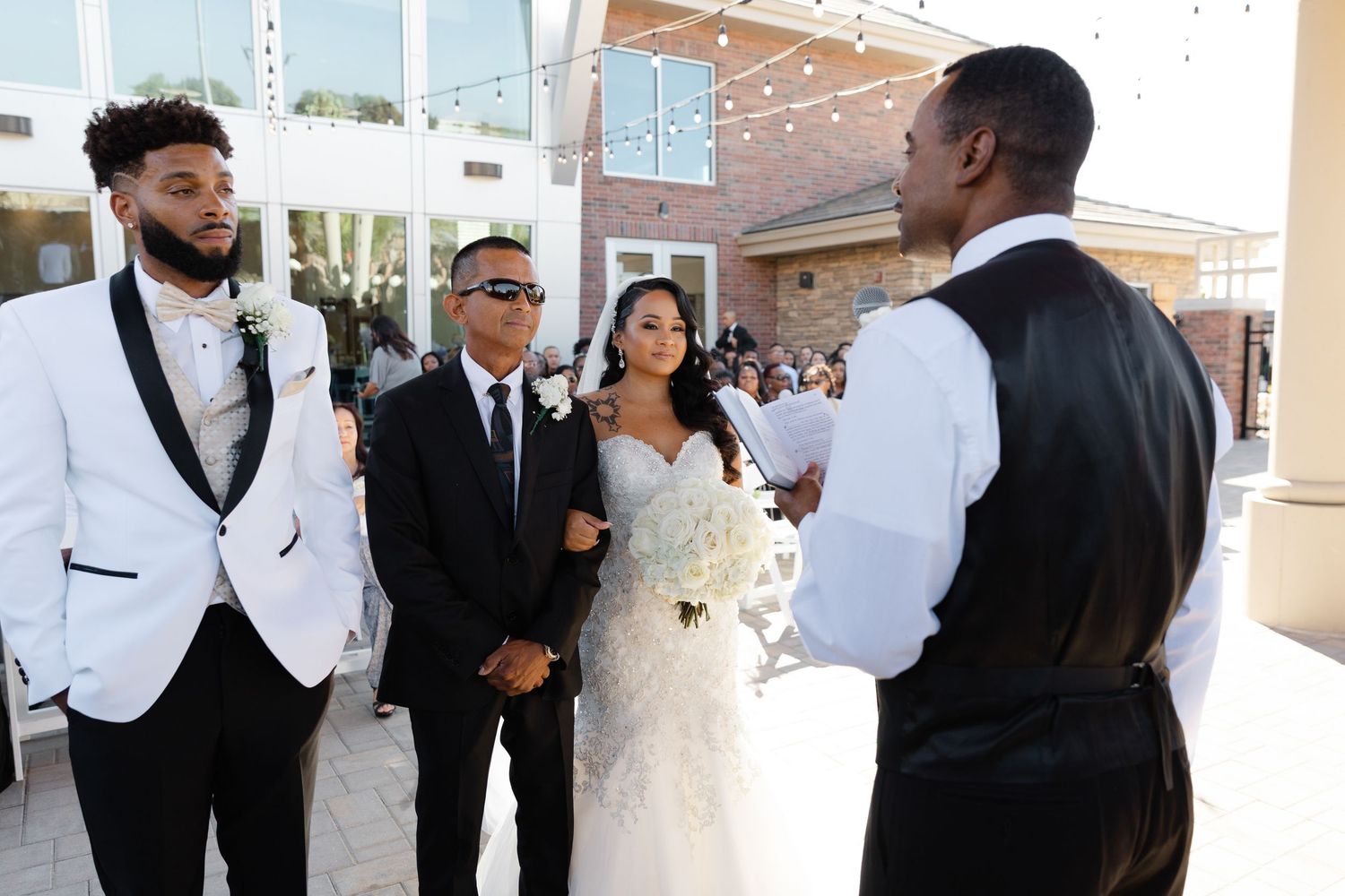 Wedding party members in formal attire gather during an outdoor ceremony with white building in background.