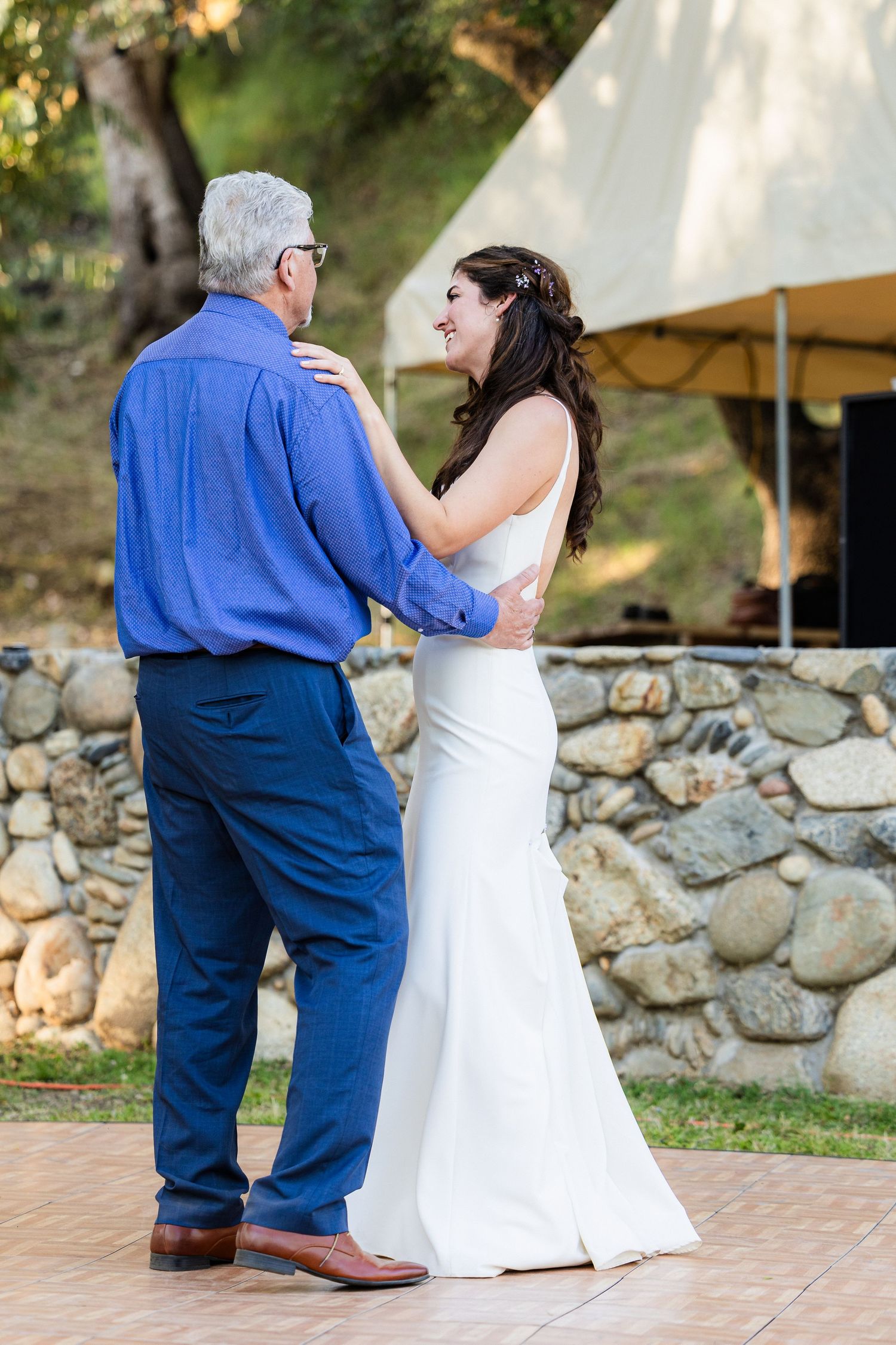 A bride in a white wedding dress shares a tender dance moment with a partner in blue at an outdoor reception.