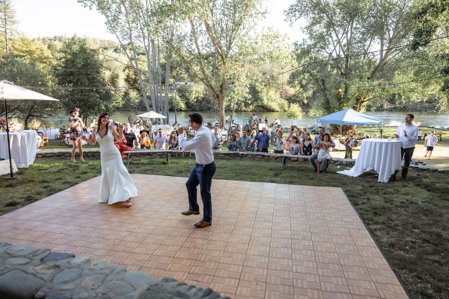 Wedding guests gather on an outdoor patio surrounded by trees during a daytime celebration.