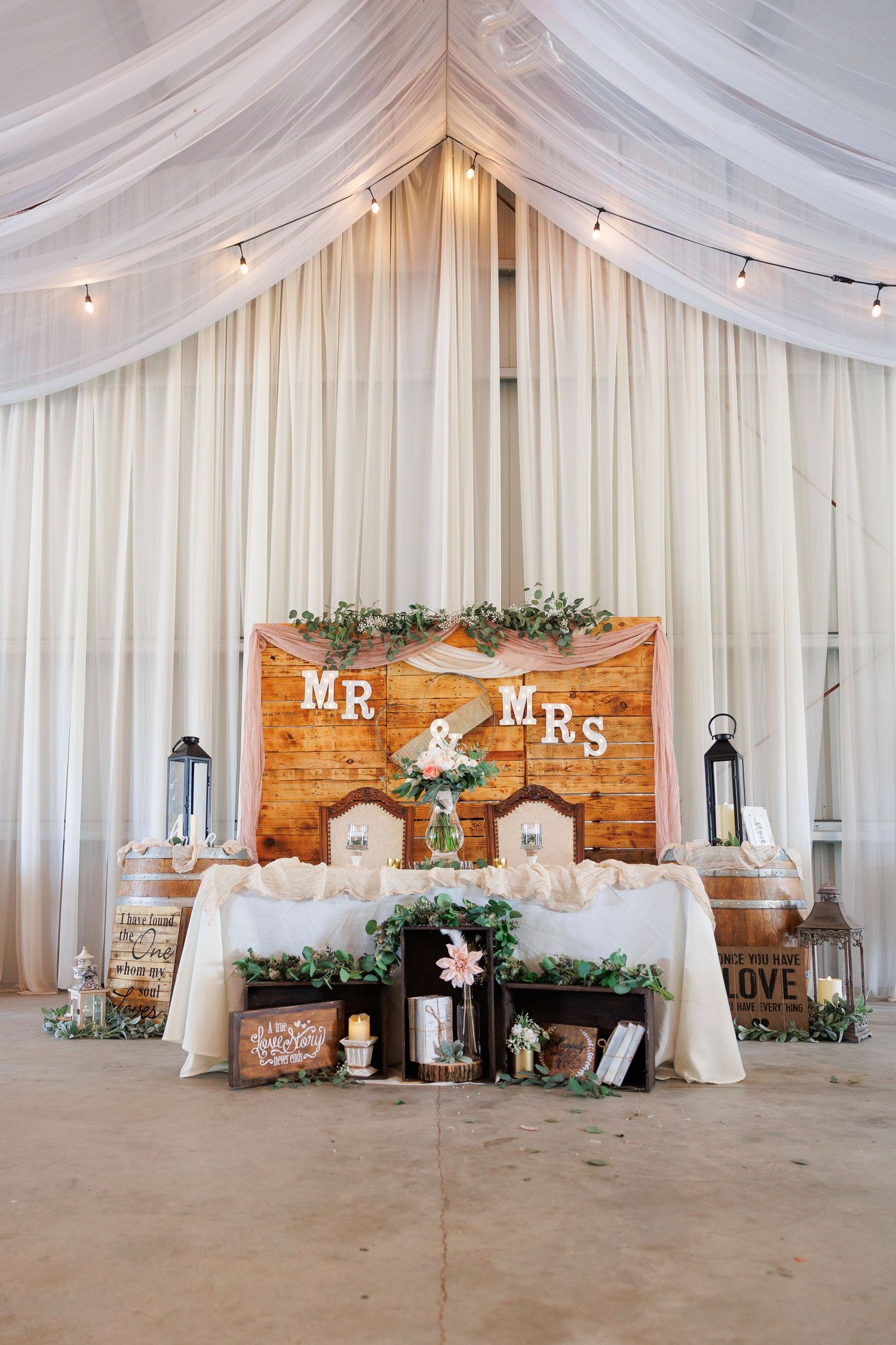Rustic wedding reception venue with white drapes, string lights, wooden backdrop, and Mr & Mrs signs decorated with greenery.