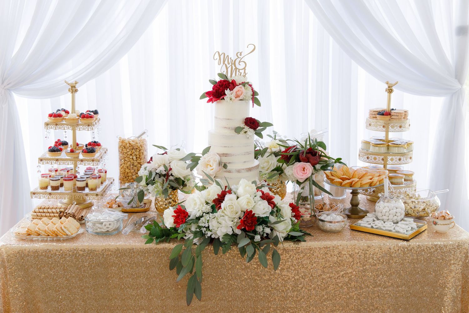 Elegant dessert table display with tiered cake stands, pastries and floral decorations against white draping backdrop.