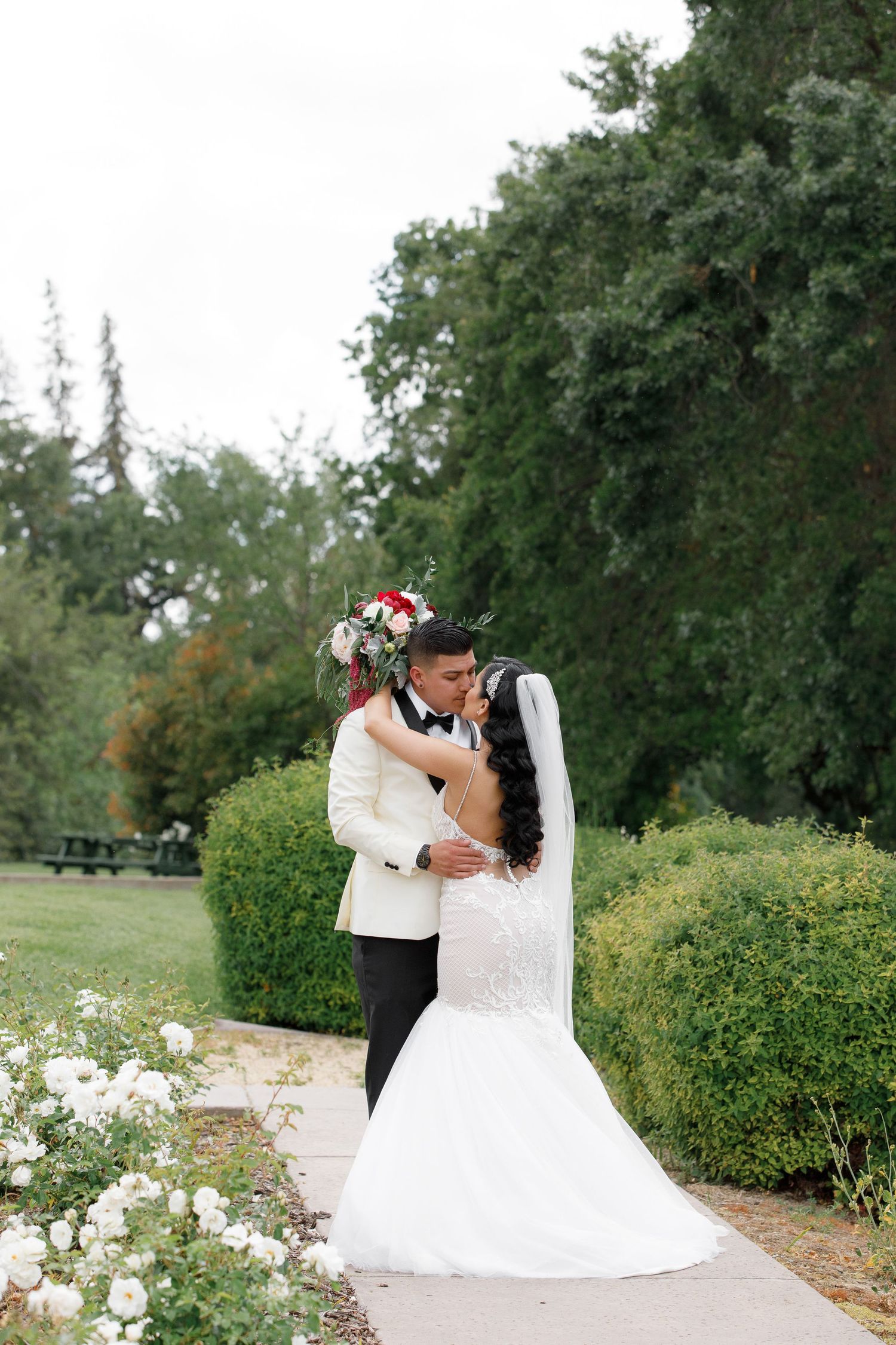 A romantic wedding couple shares tender moments in a lush garden setting with white roses and greenery.
