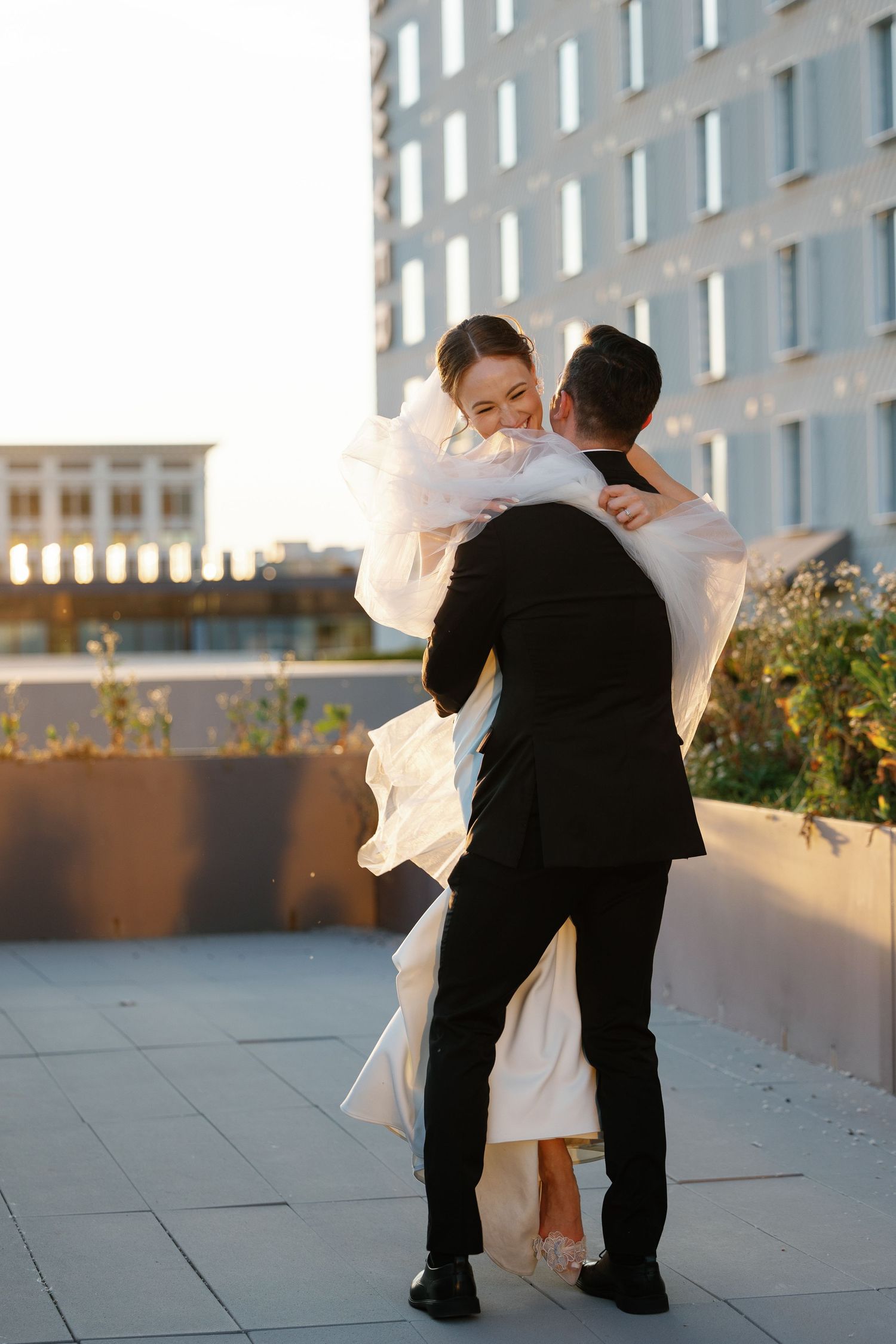 A romantic wedding couple dances together on a rooftop terrace at sunset with an urban backdrop.