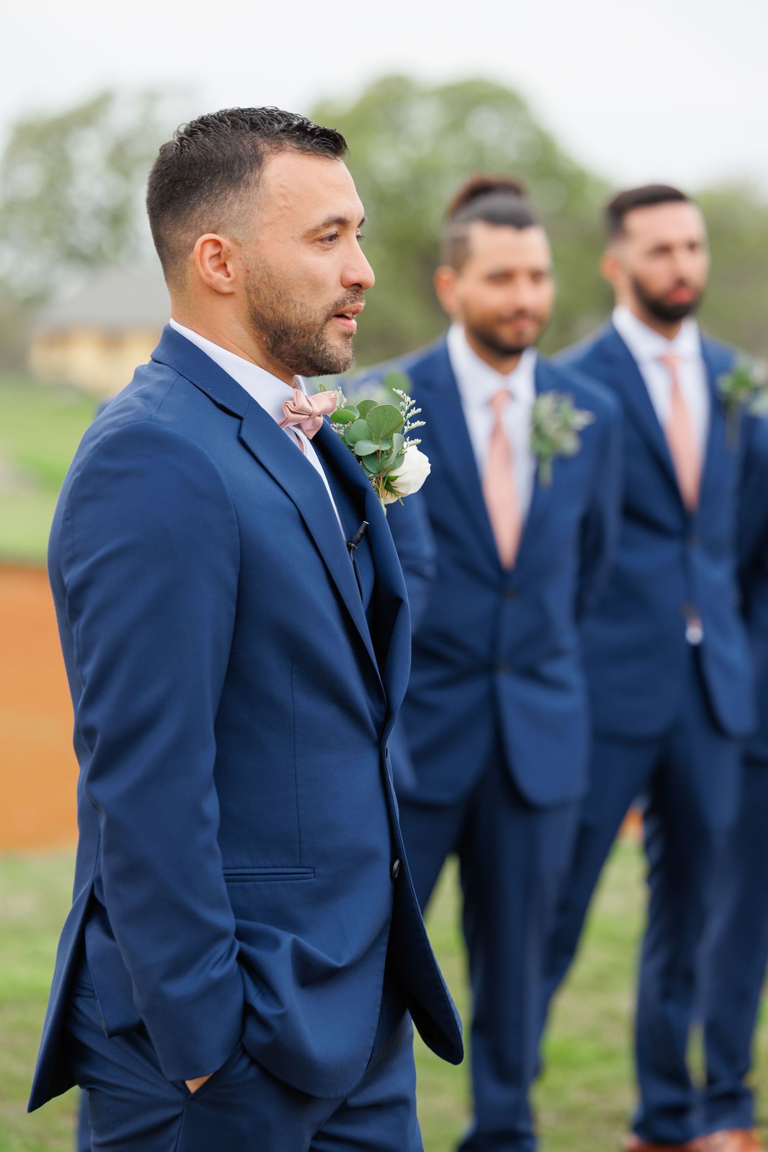 Groomsmen in navy blue suits with floral boutonnieres stand outdoors during wedding ceremony.