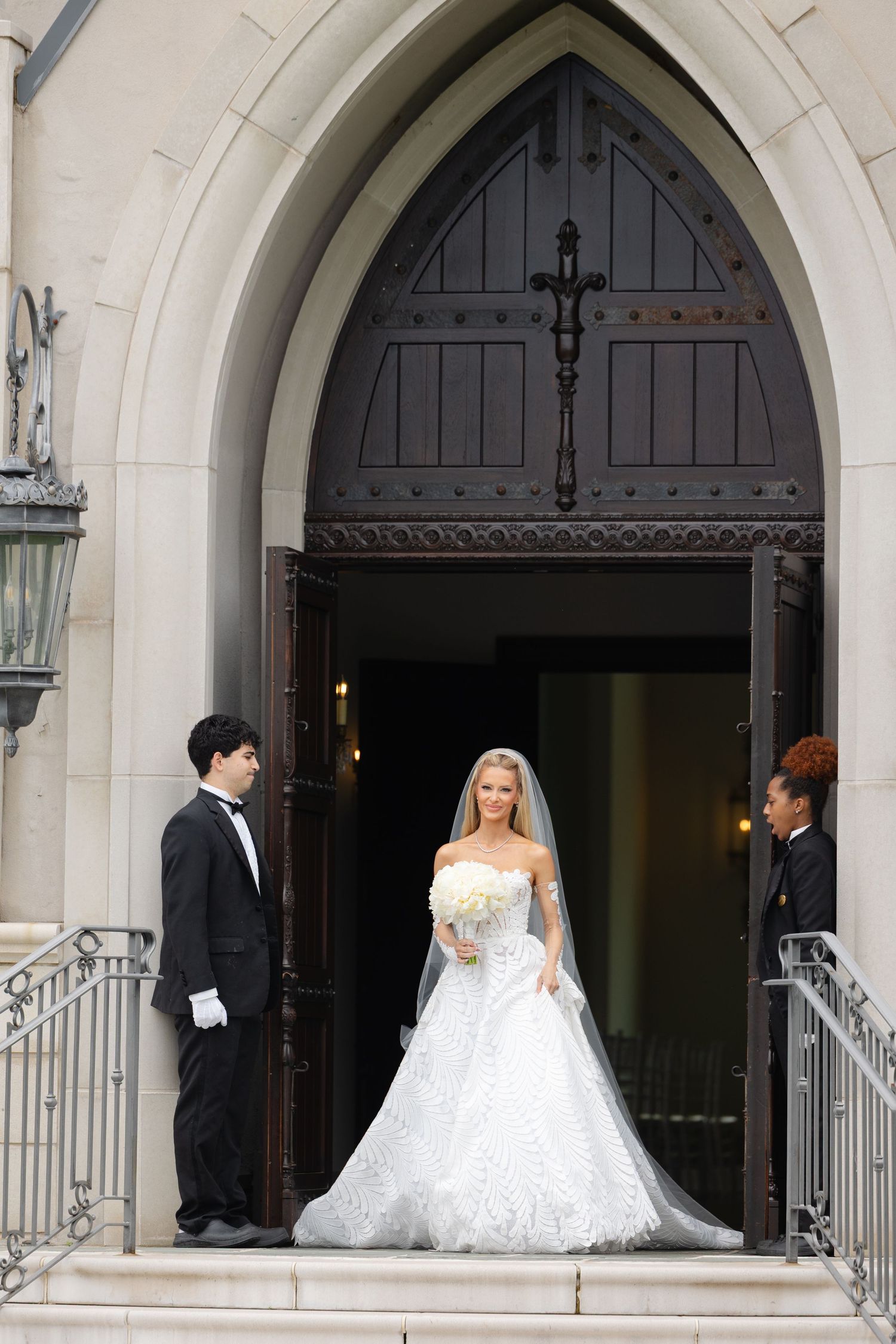 Wedding party members wait outside arched church doorways in formal attire during ceremony preparations.