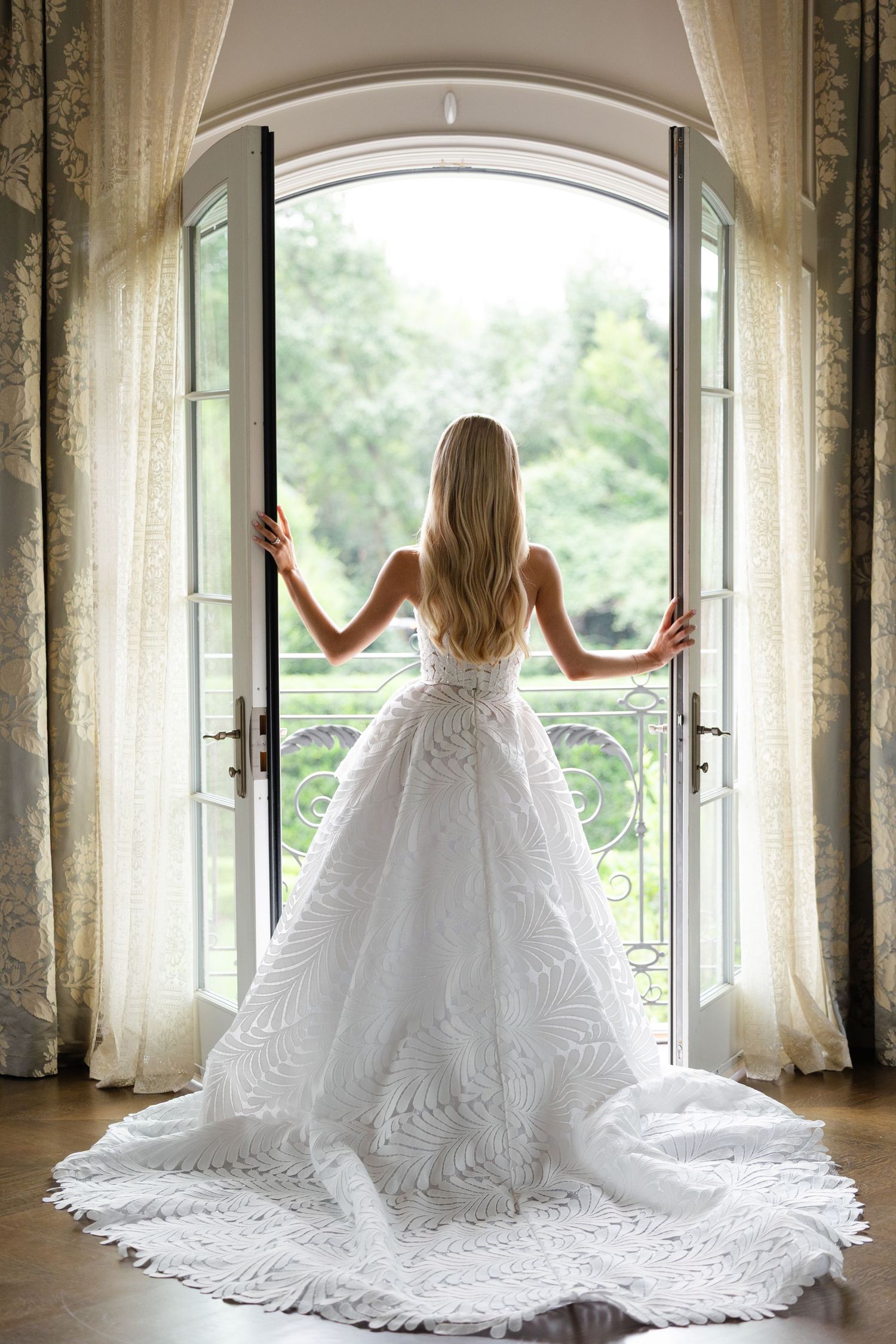 A figure in a flowing white wedding dress sits elegantly in an arched window corridor with natural light streaming in.