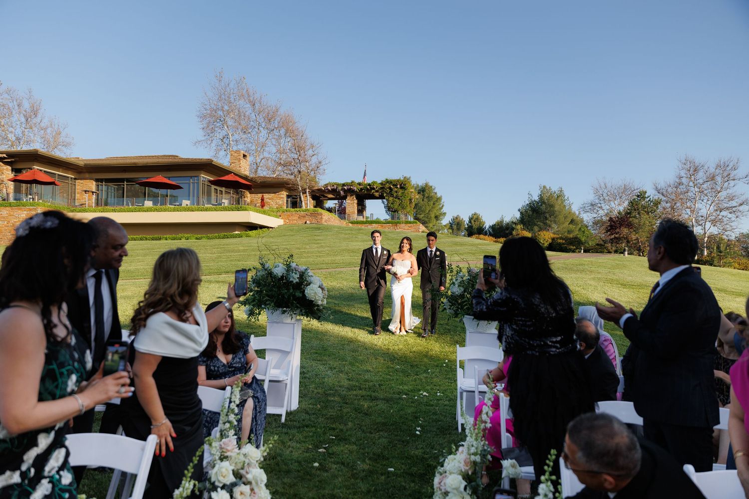 Wedding guests gather on a grassy hillside during an outdoor ceremony at sunset.
