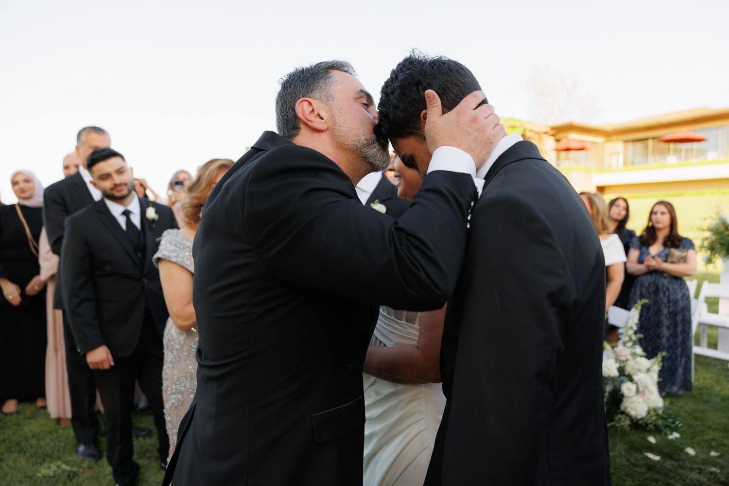 Two people in black suits share an emotional embrace at an outdoor wedding ceremony while guests look on.