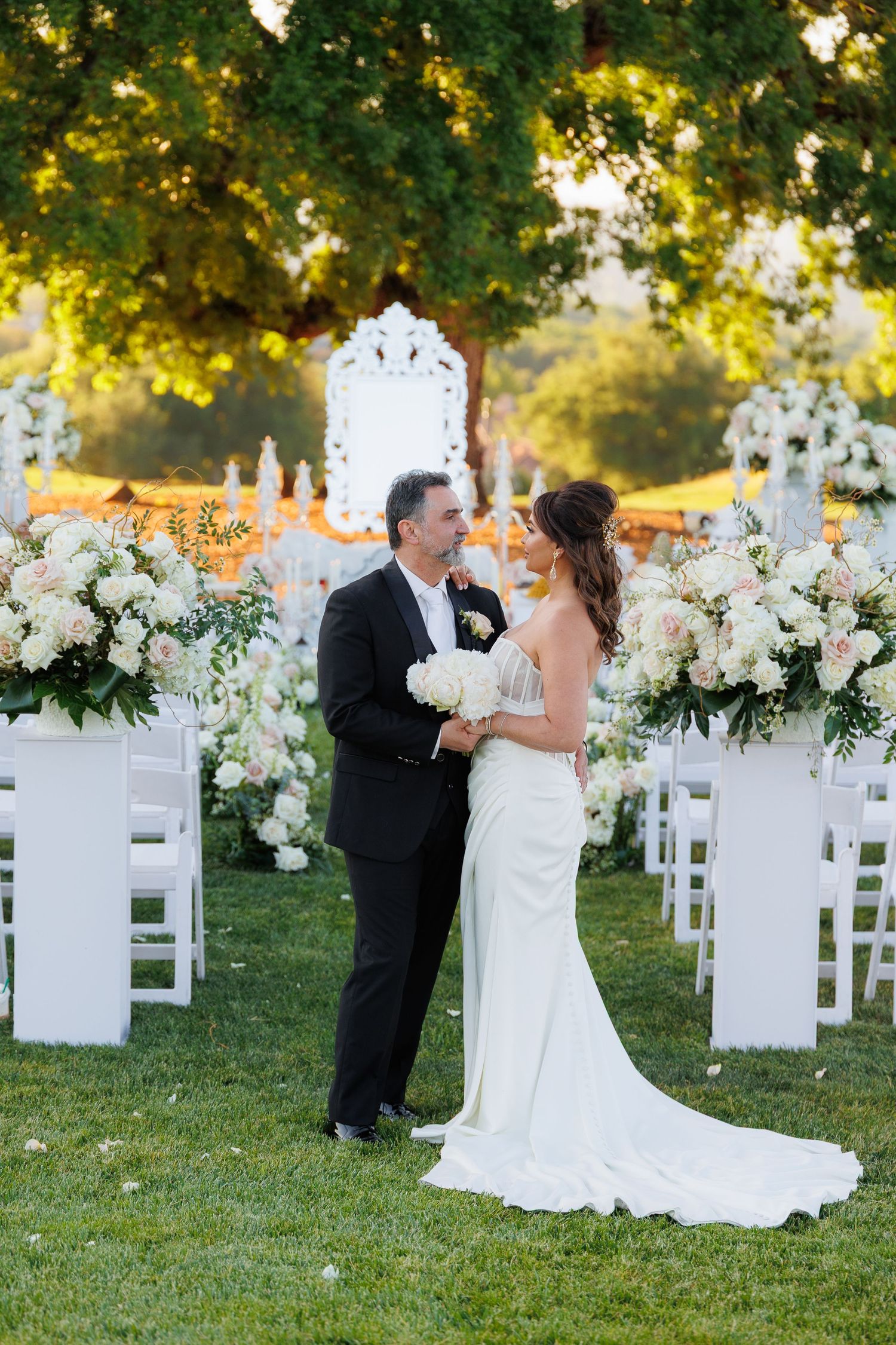 Outdoor wedding ceremony setup with white chairs and floral arrangements under trees at sunset.